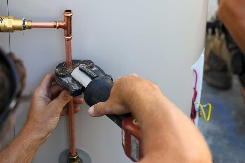 Hands using a press tool on copper pipework beside a hot water tank